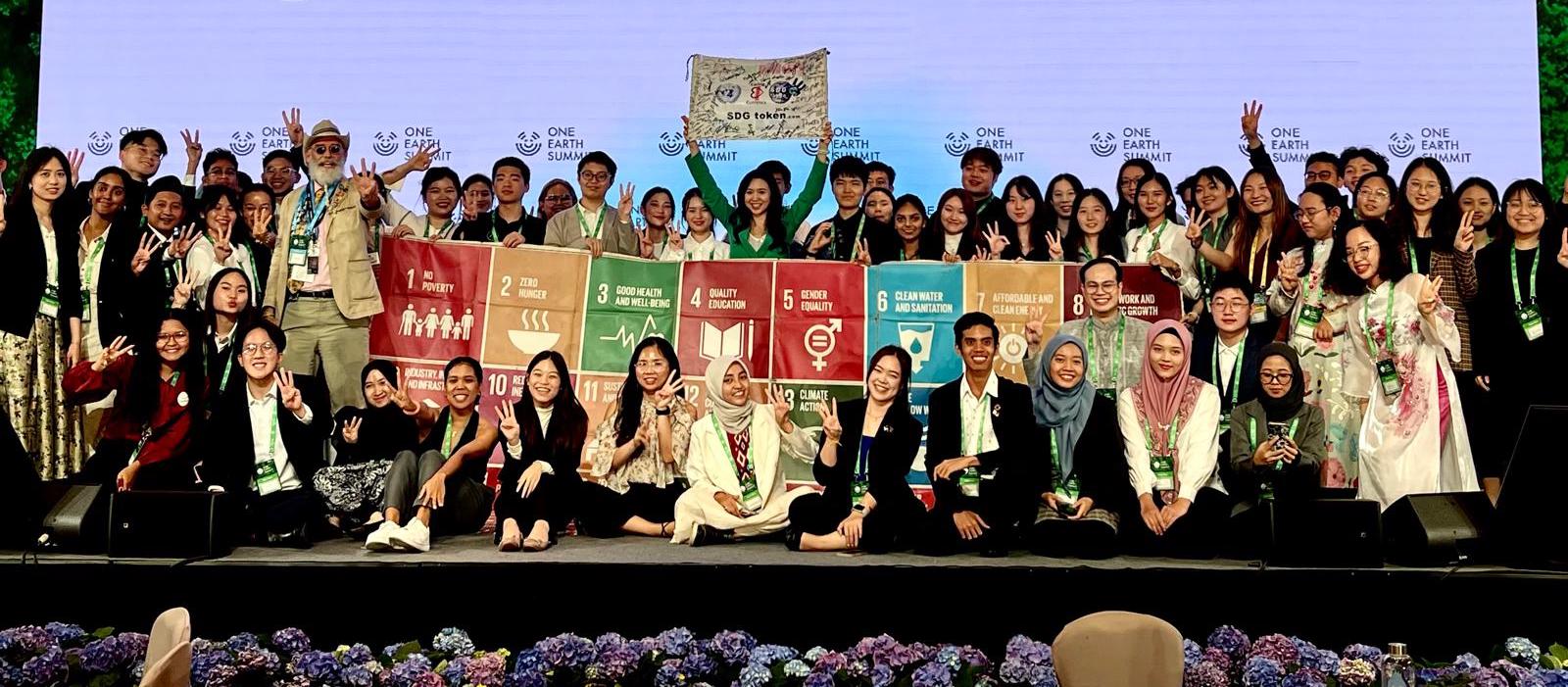 Young delegates at the One Earth Summit holding the seventeen SDG banner panels and the signed SDG token banner on stage